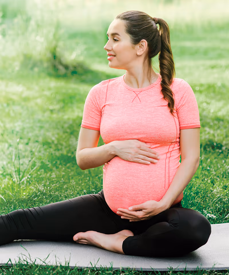 A pregnant woman with workout gear on sitting on a yoga mat outdoors in grass.