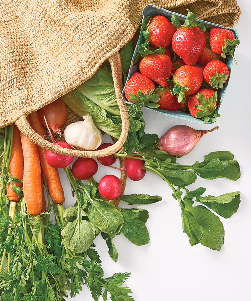 A woven market bag laying out on a surface with fresh carrots, radishes, garlic, shallots, strawberries, and herbs sticking out of it.