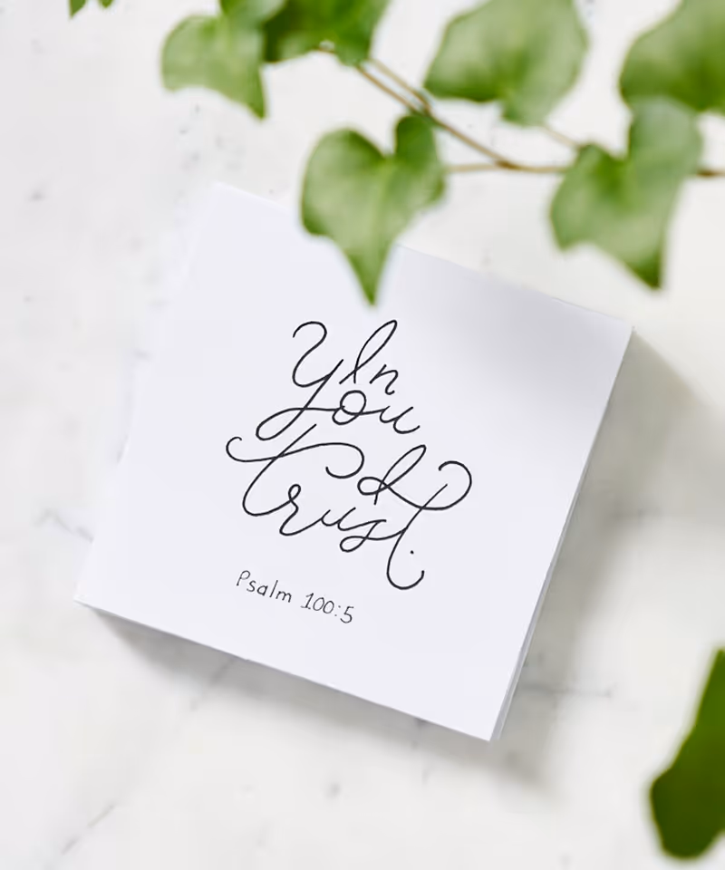 A white sticky note with the cursive text, "In You I Trust Psalm 100:5," on a white countertop with ivy.