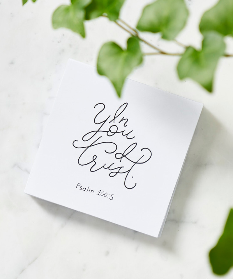 A white sticky note with the cursive text, "In You I Trust Psalm 100:5," on a white countertop with ivy.