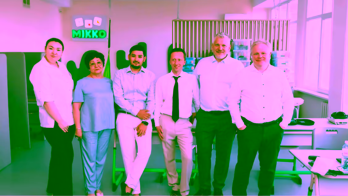 Group of six professionally dressed people standing indoors in an office setting with an open workspace background.