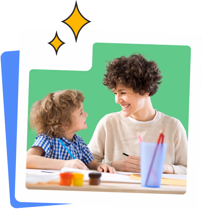 A curly-haired woman and a young boy smiling at each other while sitting at a table with art supplies.