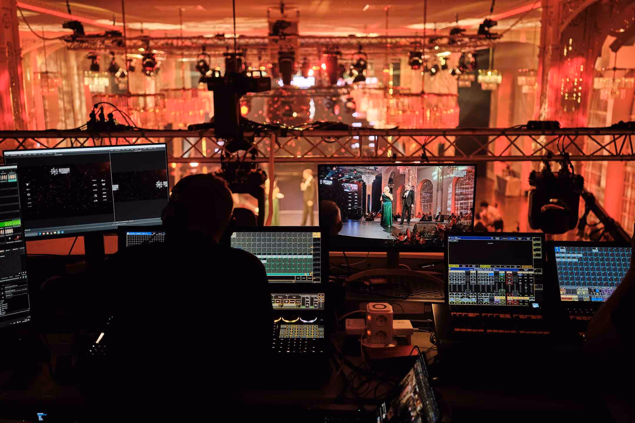 Person am Steuerpult mit mehreren Monitoren steuert Beleuchtung und audiovisuelle Effekte bei einer elegant gekleideten Gala-Veranstaltung.