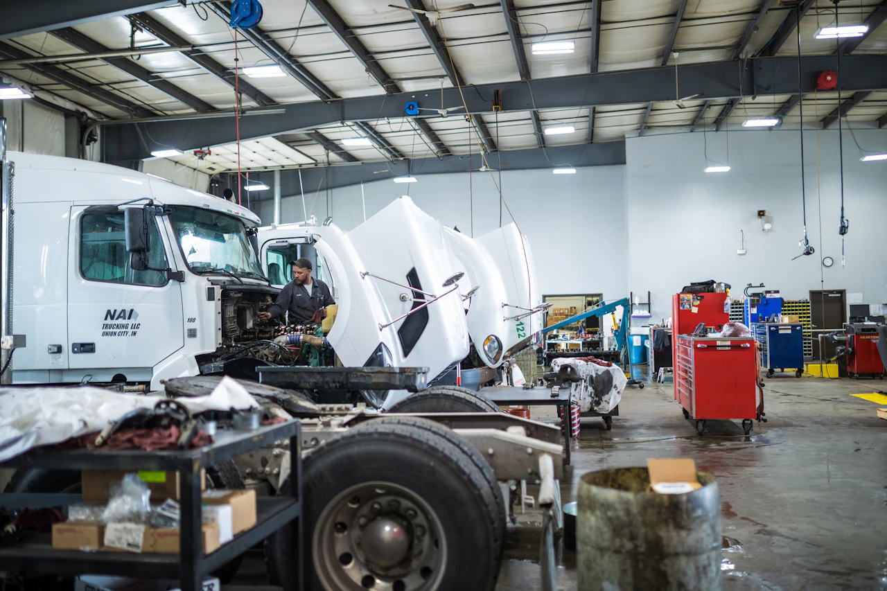 A mechanic works on the exposed engine of a white semi-truck in a spacious, well-lit garage with tools, parts, and equipment scattered around. The truck hood is lifted, revealing detailed engine components.
