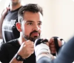 Man with dark hair and beard wearing black shirt holding boxing gloves up in a training stance.