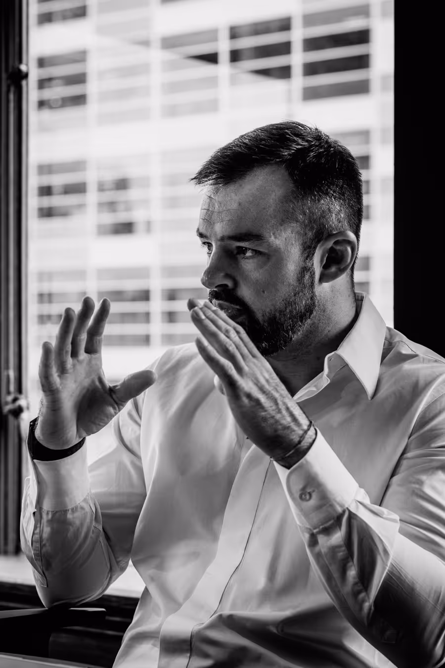 Man in white shirt gesturing with hands while speaking, sitting near a window with a building visible outside.