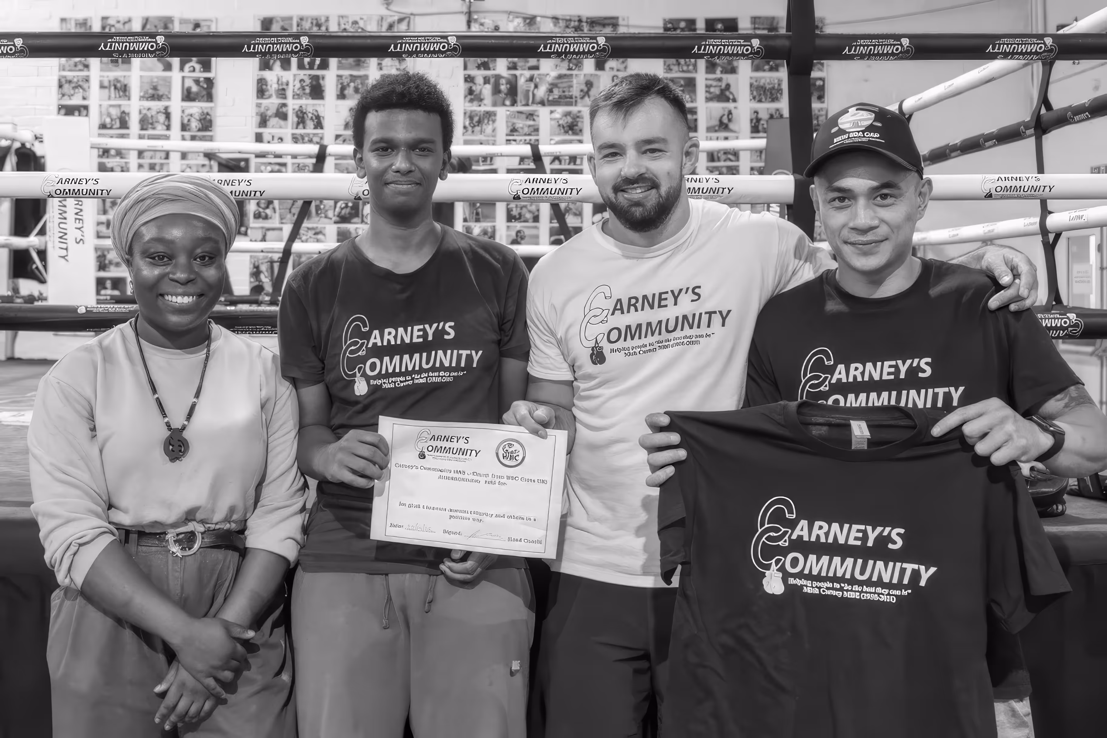 Four people standing together in a boxing gym, three wearing 'Carney's Community' shirts, one holding a certificate, another holding a T-shirt.