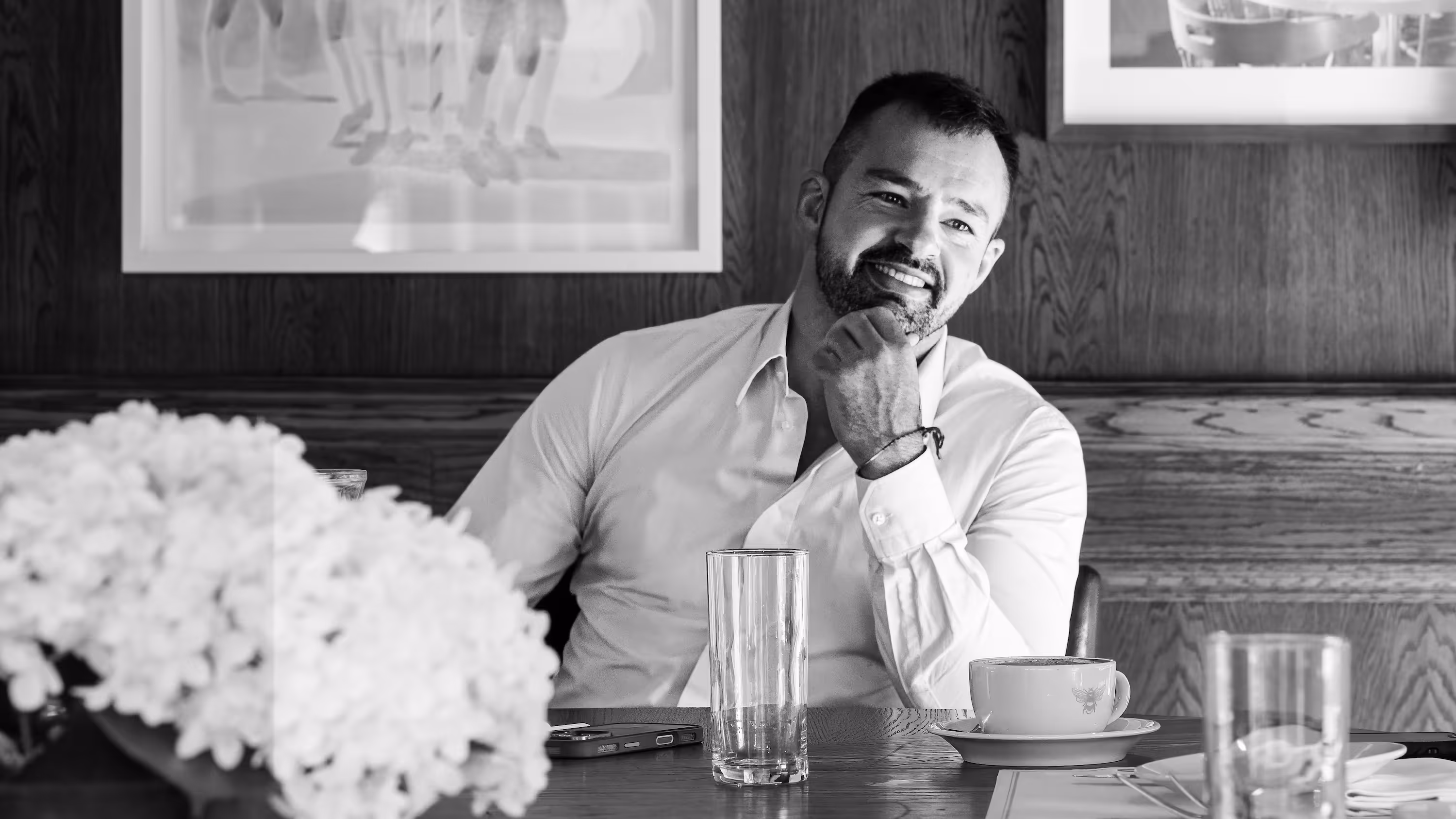 Smiling man in a white shirt sitting at a wooden table with a glass, coffee cup, and flowers in a black and white photo.