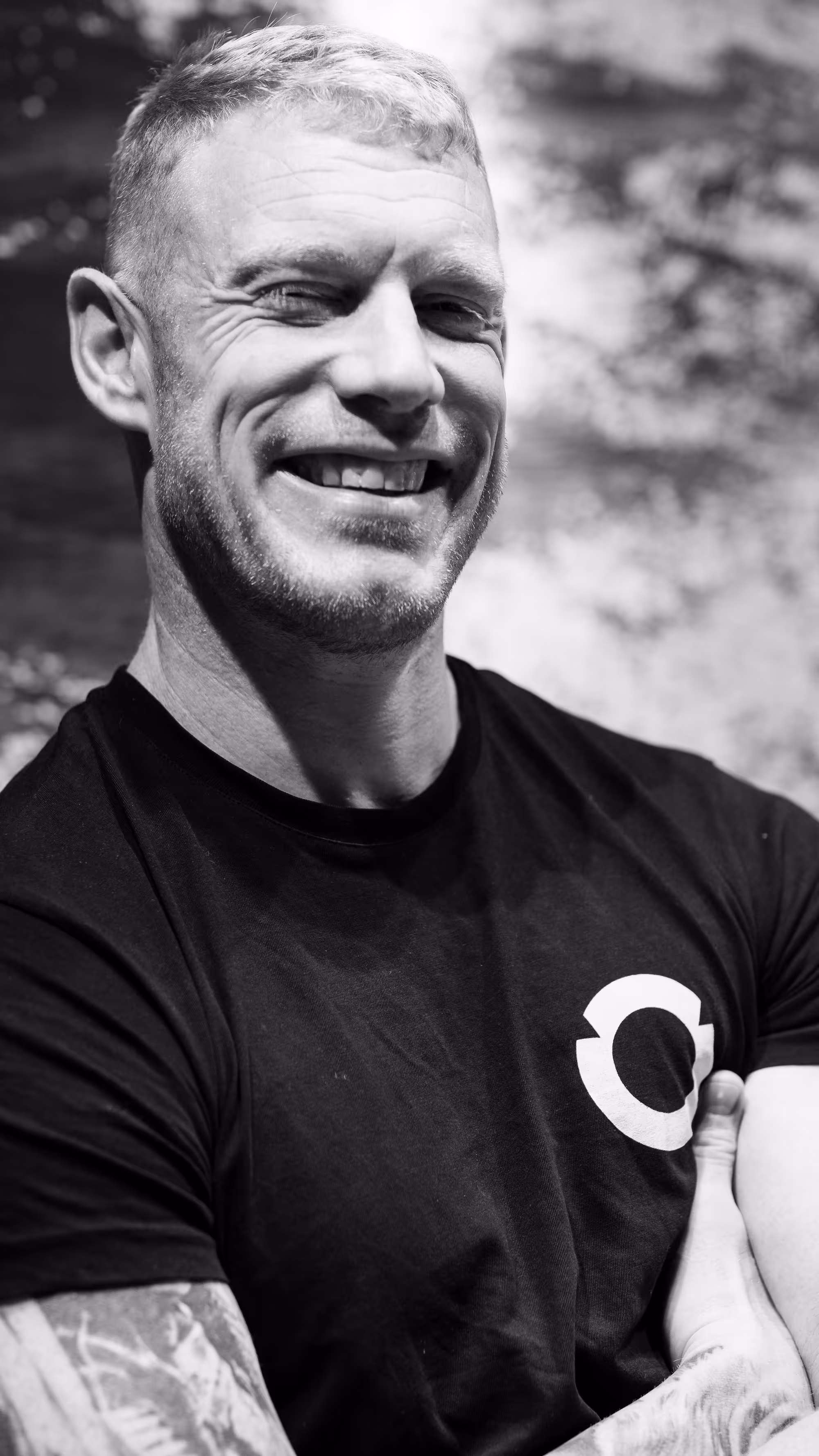 Smiling man with short blond hair, tattoos on his arm, wearing a black t-shirt with a white circular logo.
