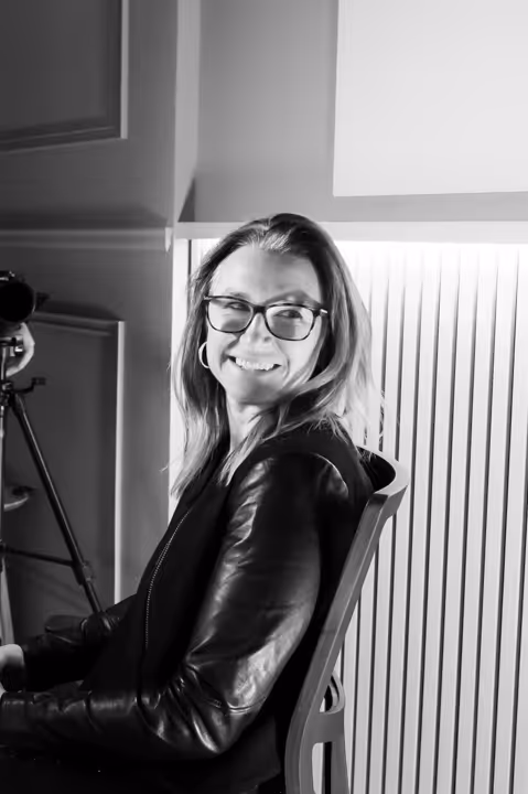 Woman wearing glasses and a leather jacket smiling and sitting on a chair in a black and white photo.