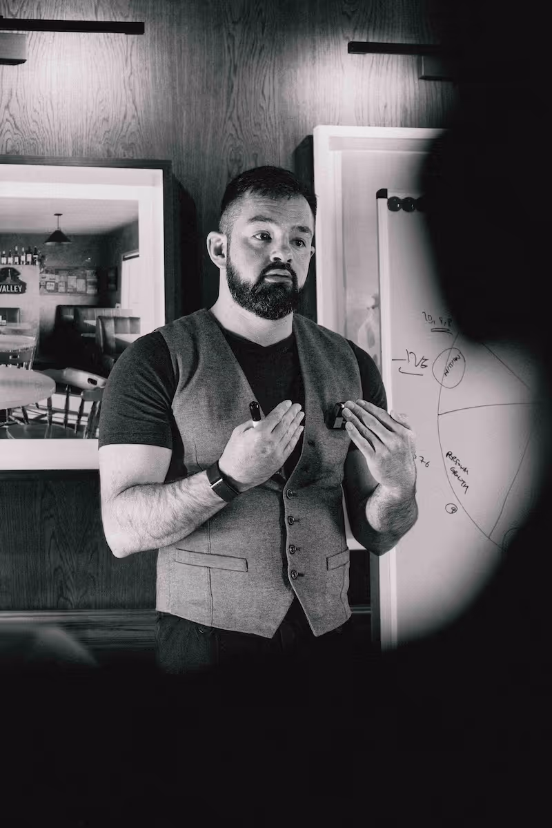 Bearded man wearing a vest speaking and gesturing in front of a whiteboard with charts in a room with wooden wall and framed pictures.