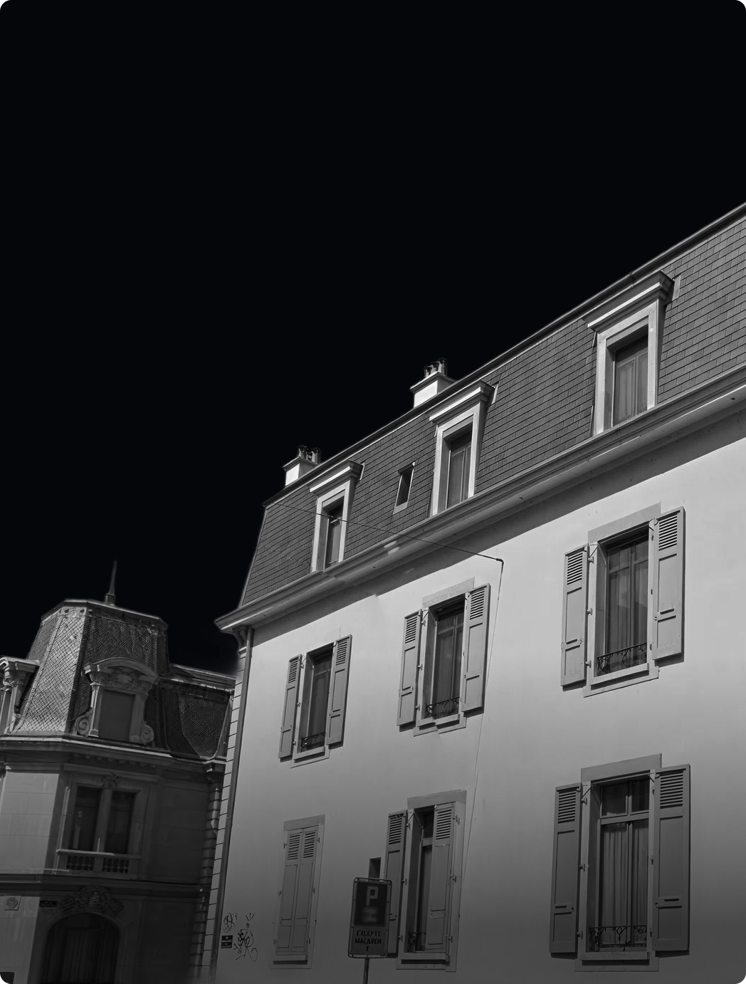 Black and white photo of a European-style building with shuttered windows and a steep roof under a dark sky.