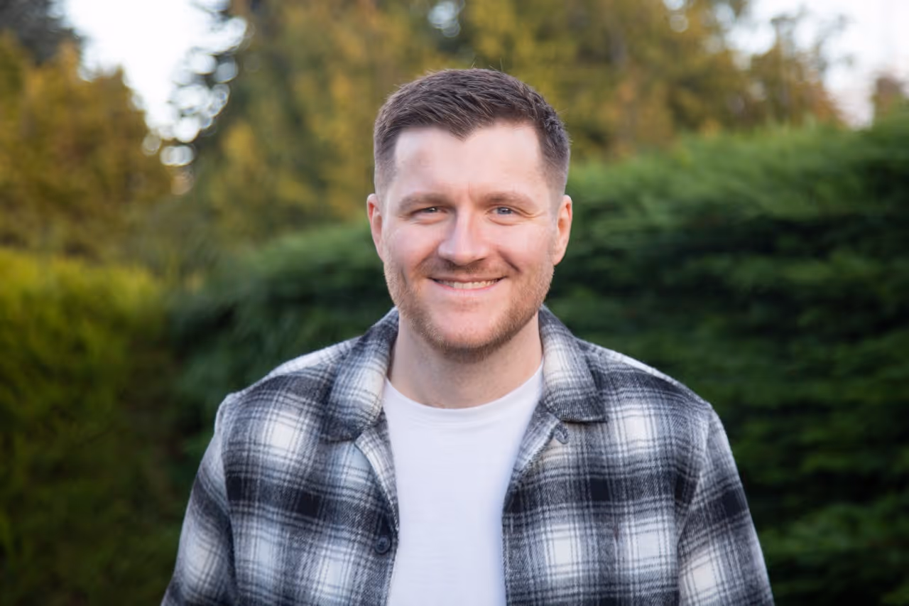Smiling person in plaid jacket standing outdoors with trees in background