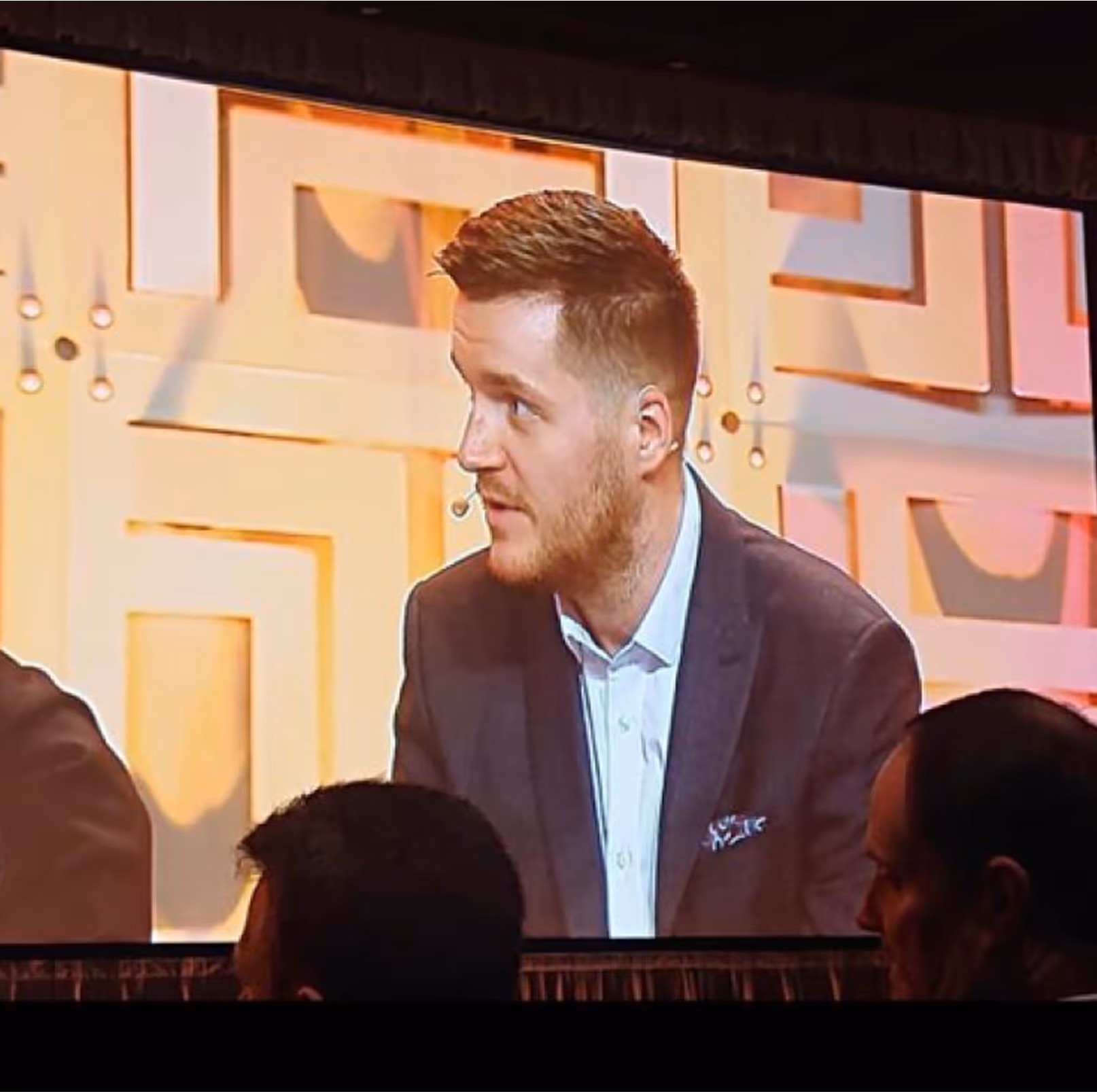 Man in brown jacket speaks on stage with geometric background