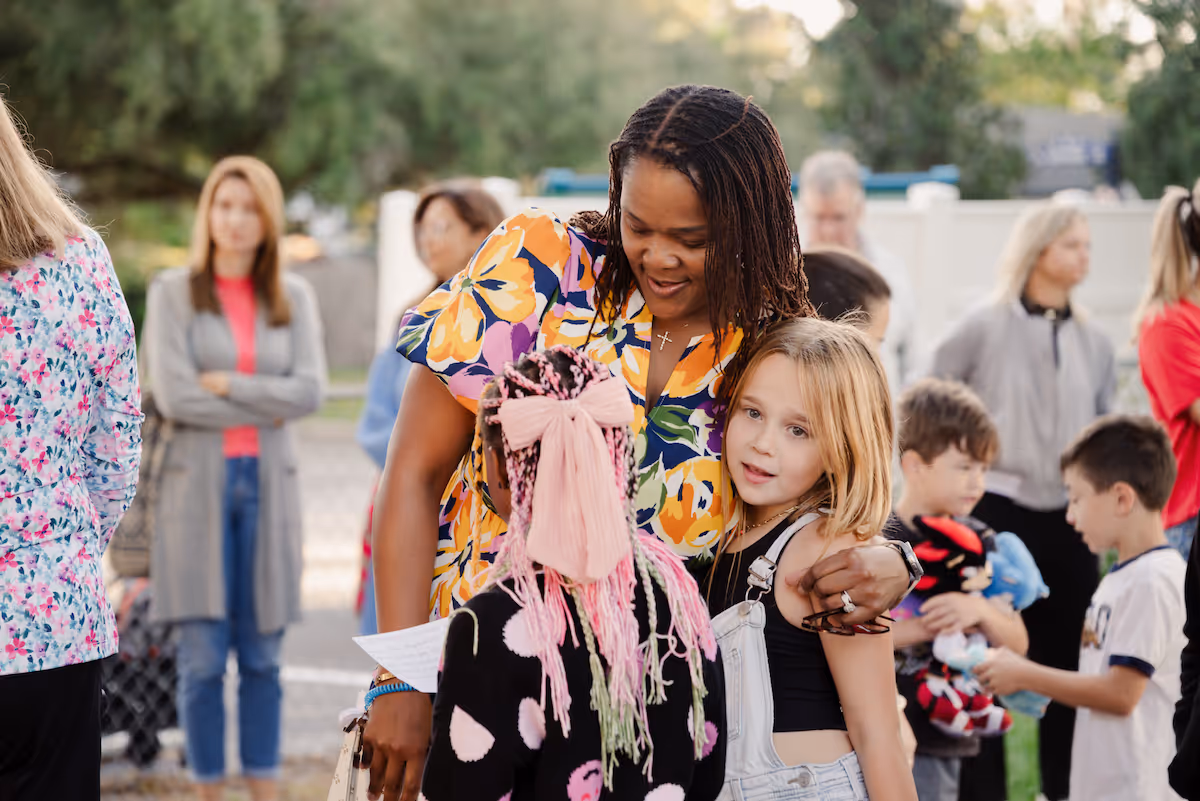 Woman in a floral dress hugging two young girls outdoors while other children and adults stand in the background.