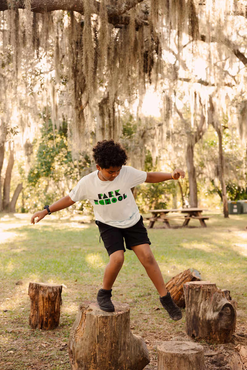 Child in a white 'Park School' t-shirt balancing on a tree stump outdoors with hanging moss and picnic tables in the background.