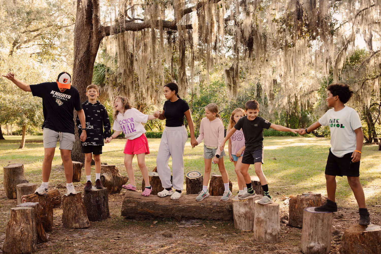 A group of children holding hands while balancing on tree stumps and a log in a wooded park area.