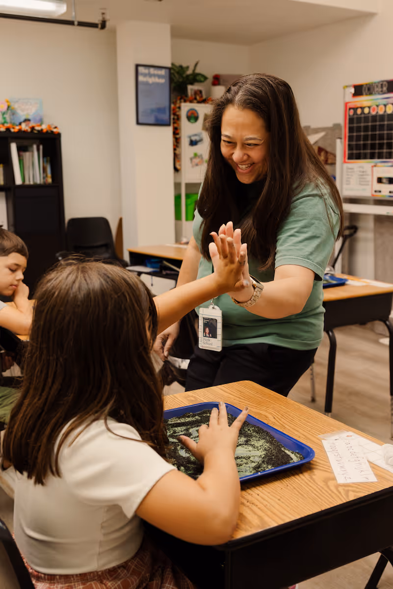 Teacher smiling and giving a high-five to a student sitting at a desk with a tray of sand.