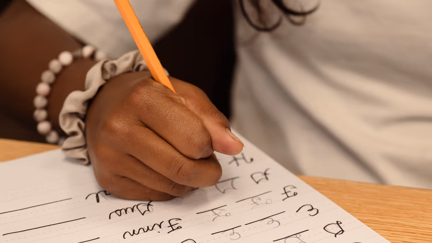 Close-up of a person writing cursive letters with an orange pencil on lined paper.