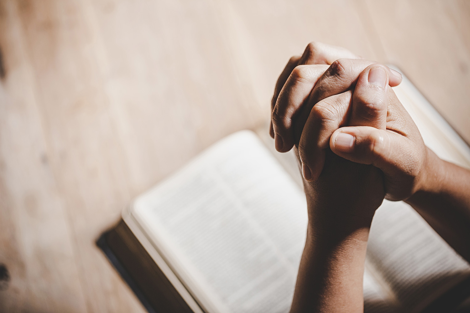 Hands clasped together in prayer over an open Bible on a wooden table.