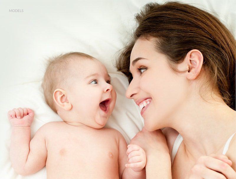 A smiling woman and happy baby lying down on a white sheet and looking at each other.