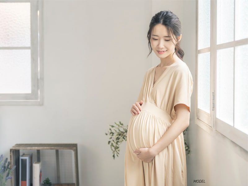 A pregnant woman in an ivory dress standing near a window.