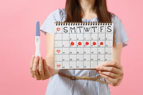happy woman in blue dress, hat hold in hand pregnancy test, periods calendar for checking menstruation days-img-blog