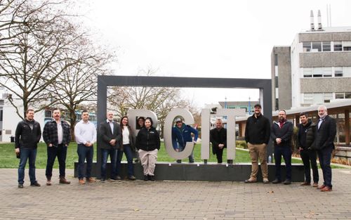 Group of people standing and posing around large metallic letters spelling 'BCIT' area near buildings.