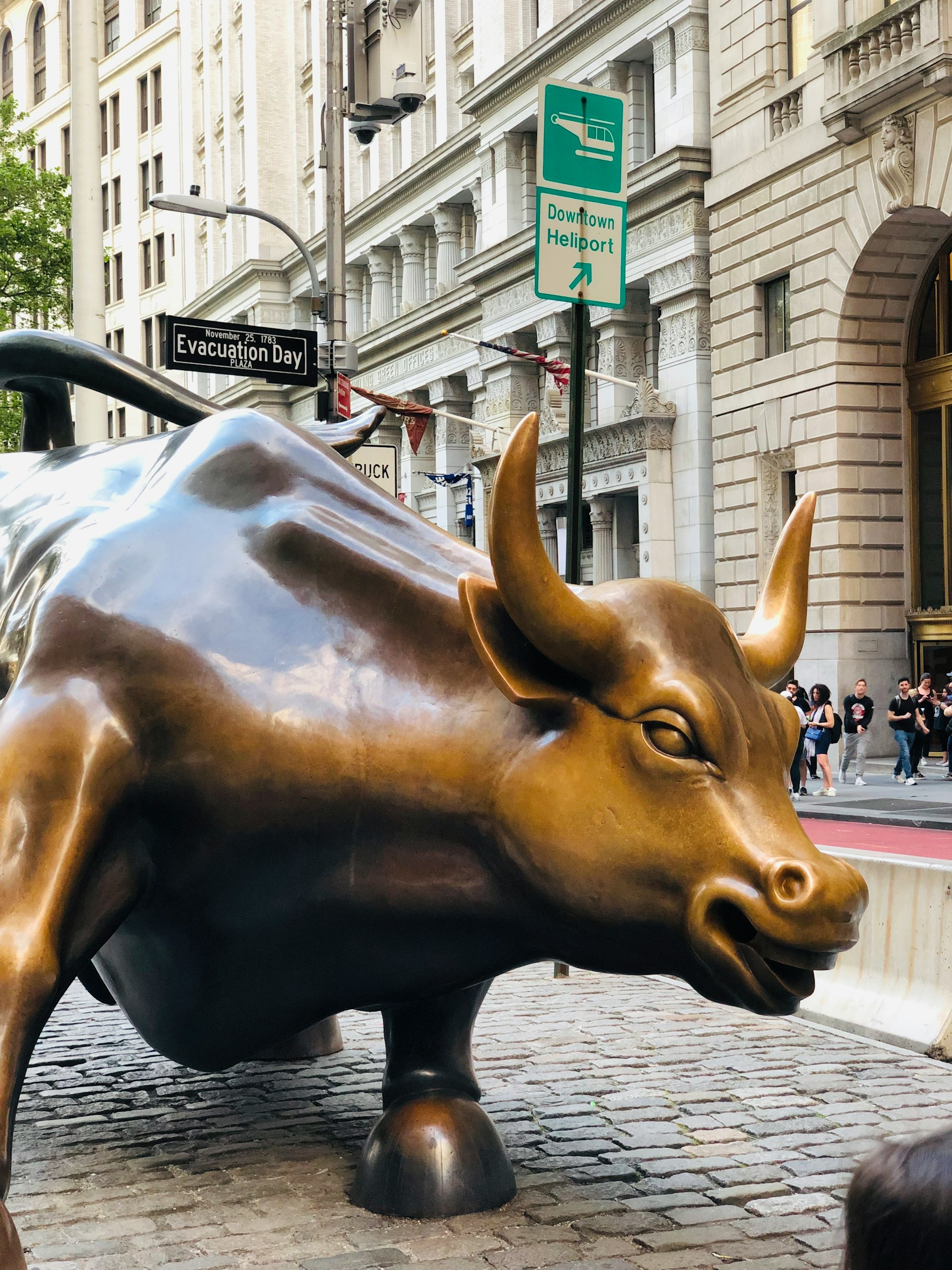 Close-up of the Charging Bull bronze statue on a cobblestone street with historic buildings and people in the background.