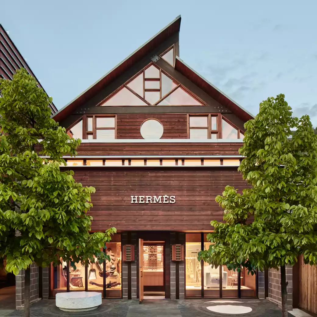 Hermès store with wooden facade and glass windows framed by green trees under clear sky.
