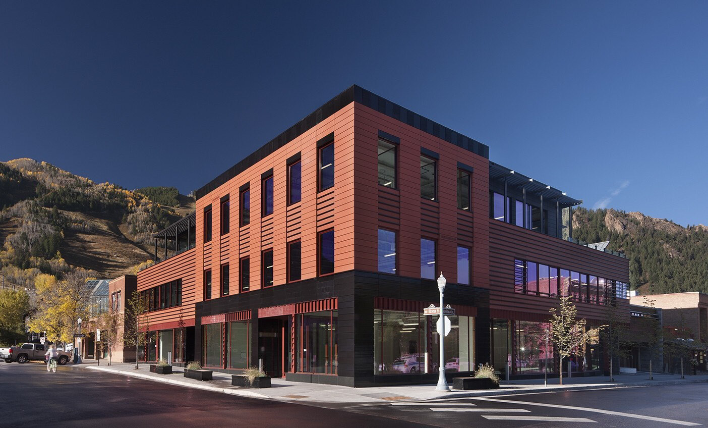 Modern two-story building with red and black exterior on a street corner, set against a backdrop of forested mountains under a clear blue sky.