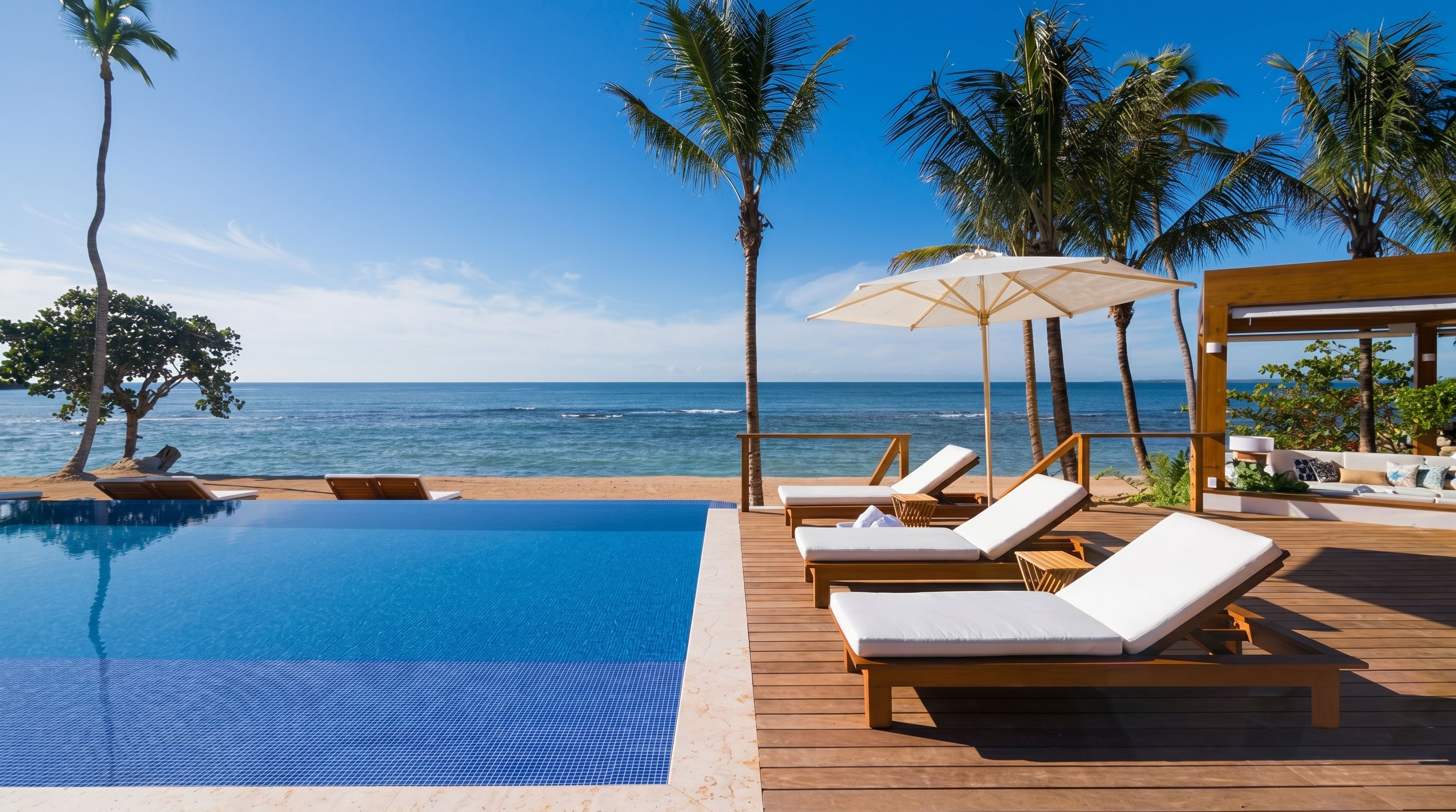 Infinity pool and teak sun loungers on a wooden deck facing the Caribbean Sea, framed by palm trees under a clear blue sky in the Dominican Republic.