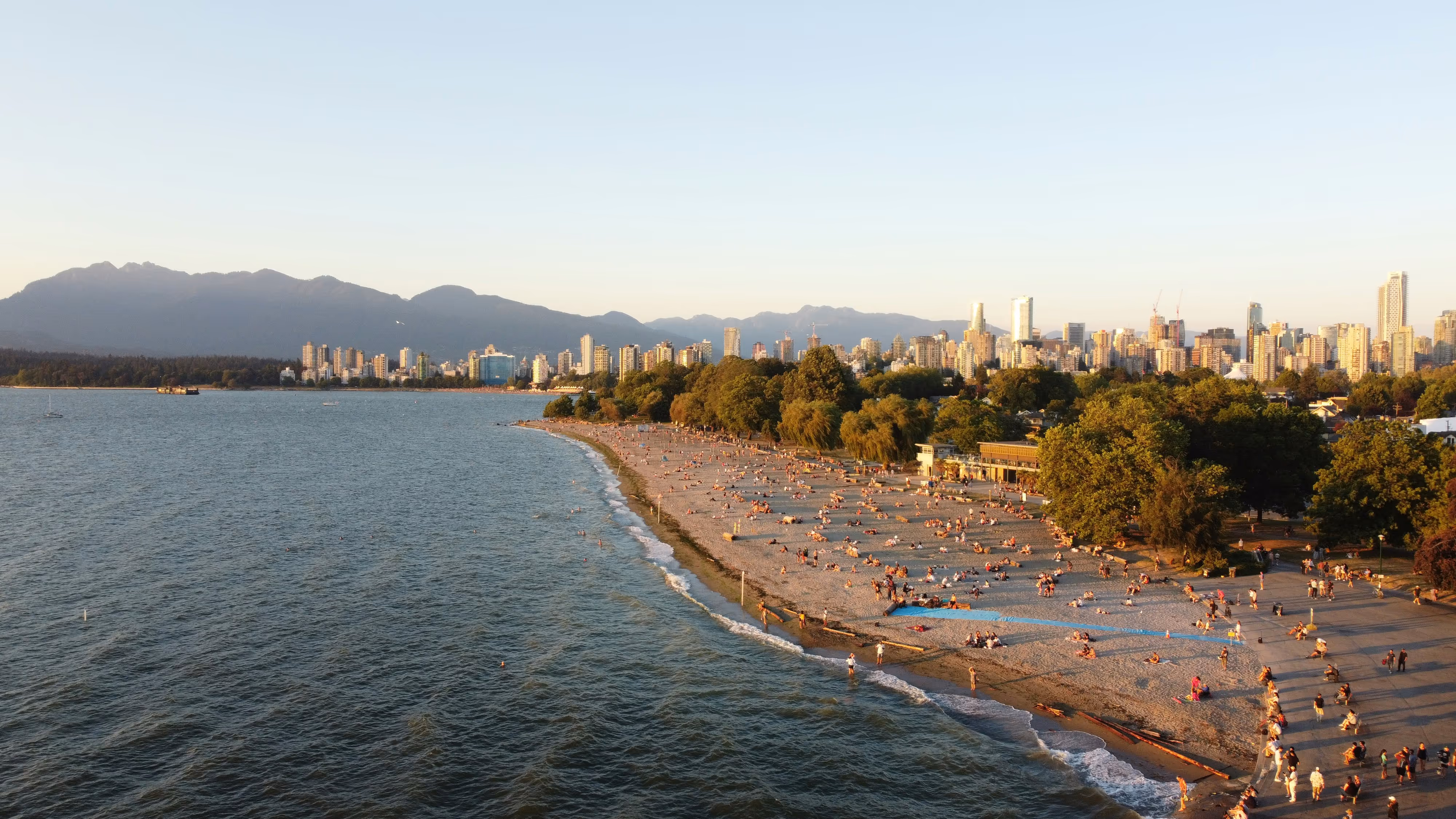 A crowded sandy beach with people relaxing by the water, green trees, and a city skyline with mountains in the background during sunset.