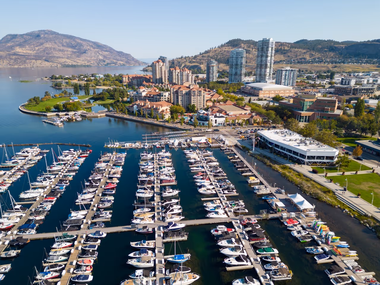 Aerial view of a marina with numerous docked boats, adjacent parkland, residential buildings, and hills in the background under a clear sky.