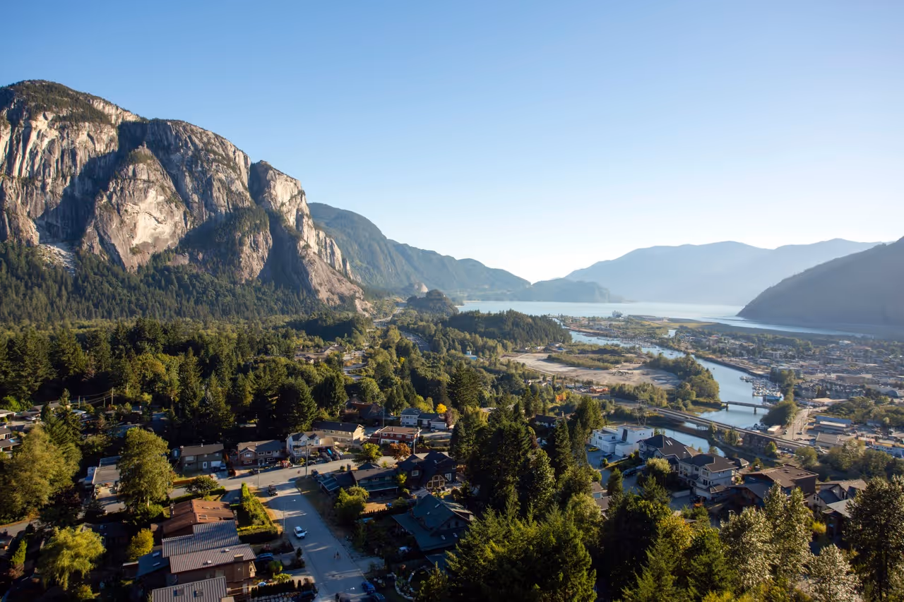 Aerial view of a town surrounded by dense trees, mountains, and a river under a clear blue sky.