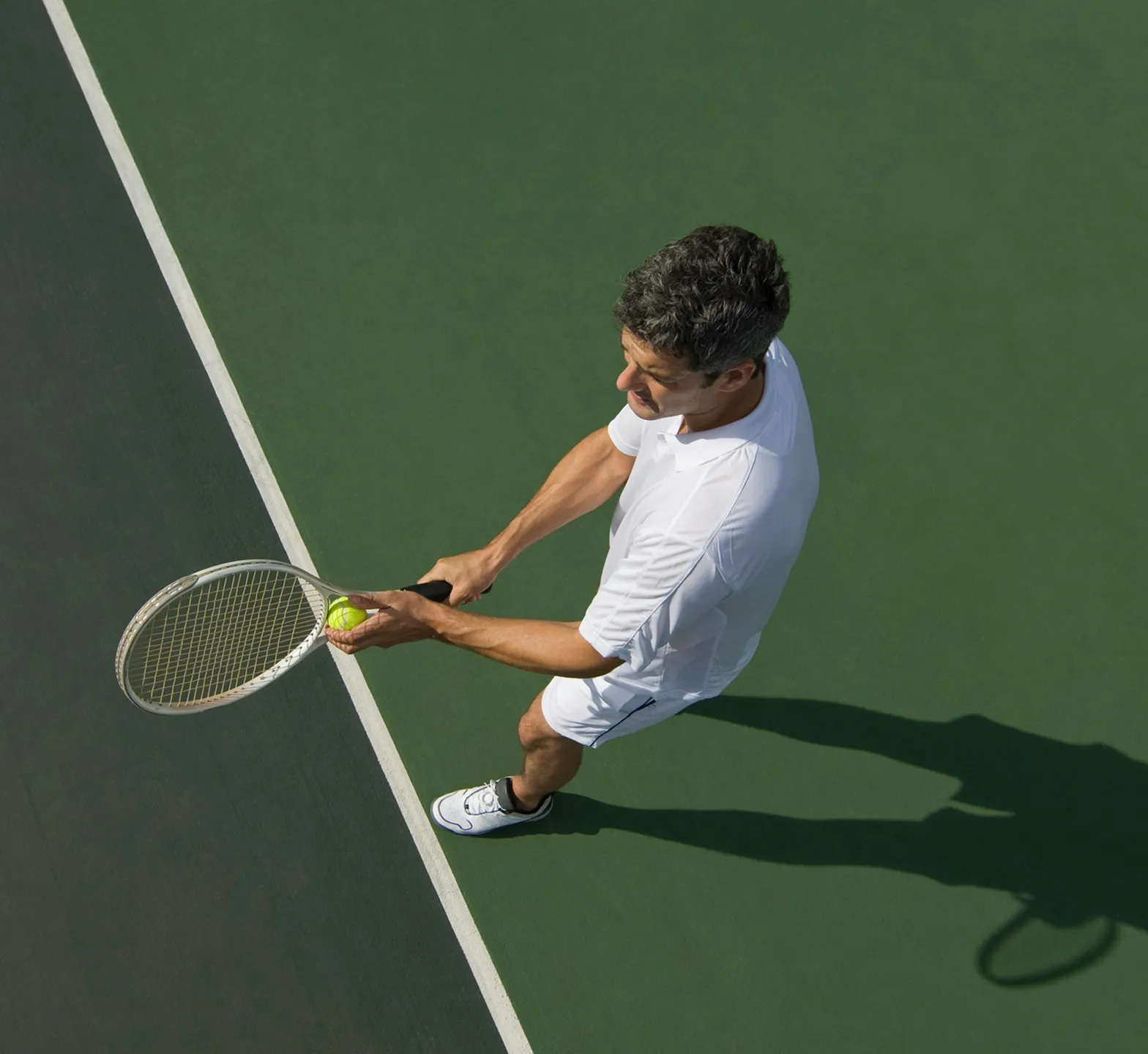 High-angle view of a tennis player in white sportswear preparing to serve on a green court