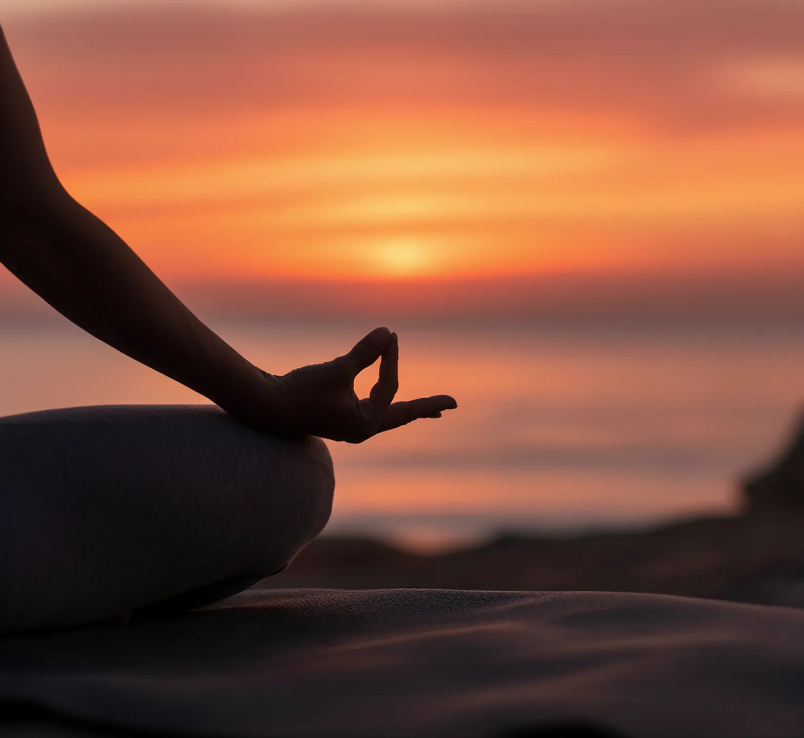 Close-up of a hand in a meditation pose against a vibrant orange sunset over the sea