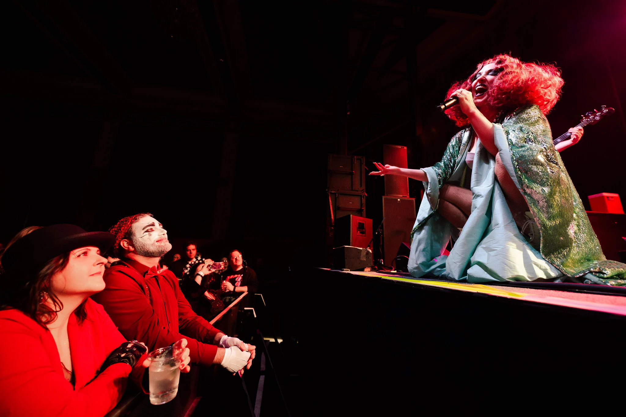 A singer in a green kimono performs from the stage edge as fans in face paint watch from the front row