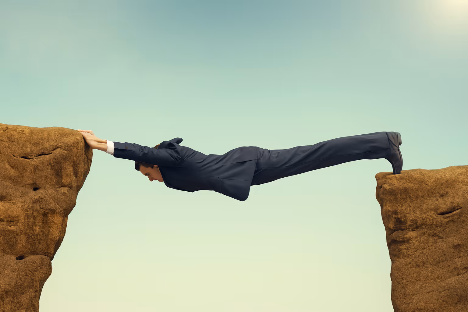Businessman in a suit stretched horizontally across a gap between two cliffs, using his hands and feet to bridge the chasm, symbolizing overcoming challenges and bridging gaps in business.