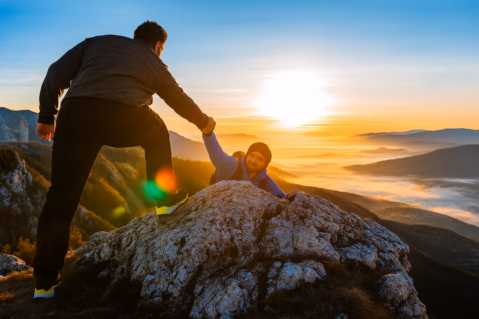 Photo of a man helping another climber up a rocky mountain summit at sunrise, representing trust, support, and collaboration.