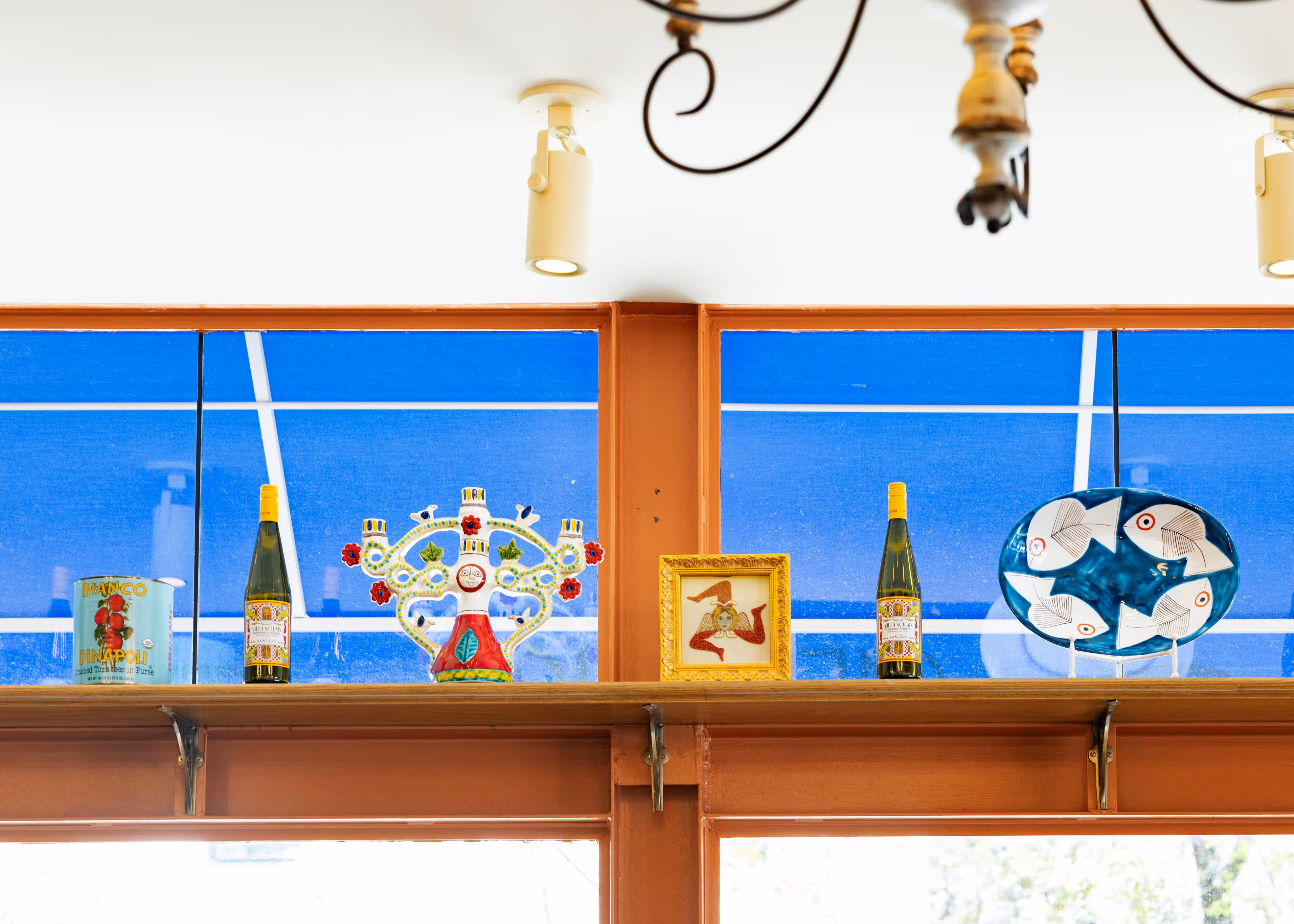 Shelf displaying two bottles of wine, a decorative ceramic piece with red flowers and faces, a framed mosaic of a figure with three legs, and a blue plate with white fish designs.