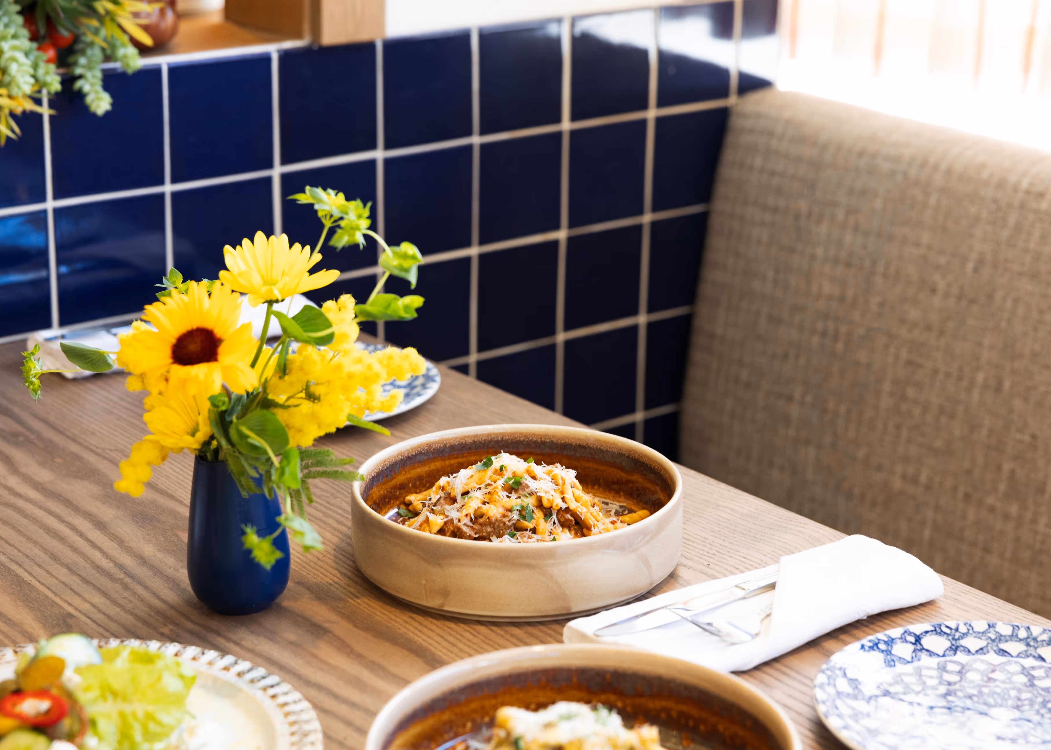 Table setting with bowls of pasta garnished with herbs, a blue vase holding yellow flowers, and a folded napkin with cutlery on the side.