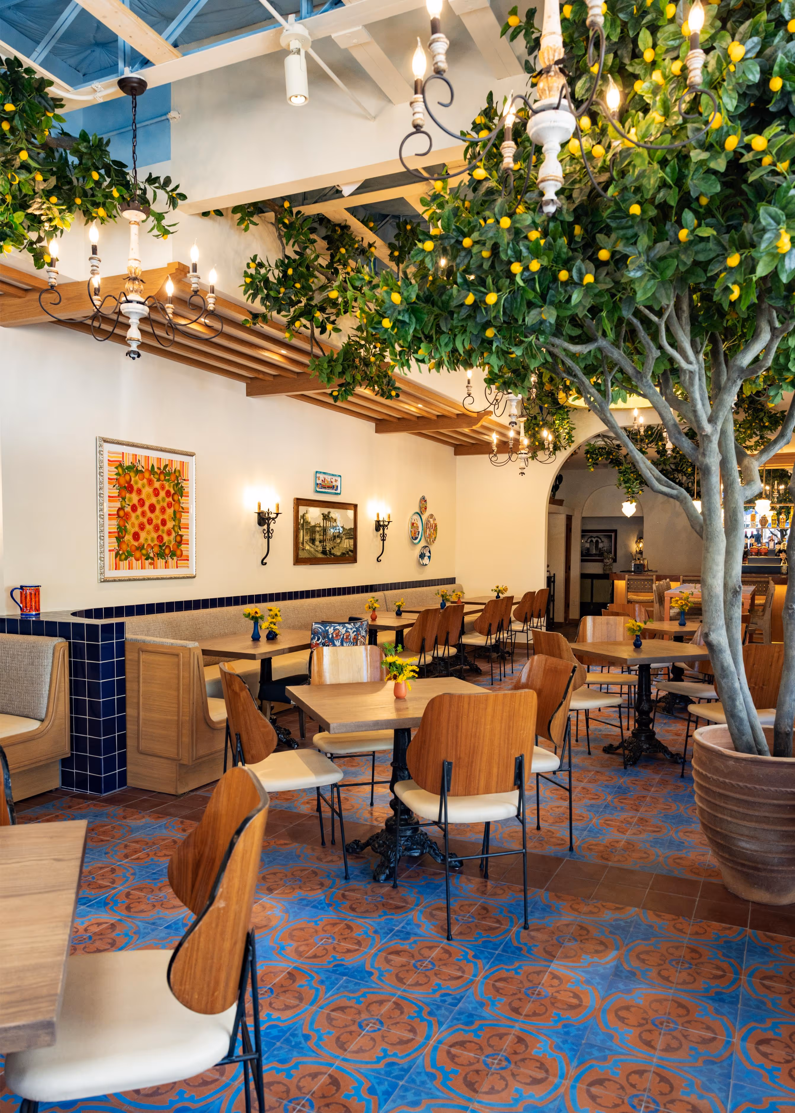 Bright restaurant interior with wooden tables and chairs, blue and orange patterned tile floor, lemon trees, chandeliers, and wall art.