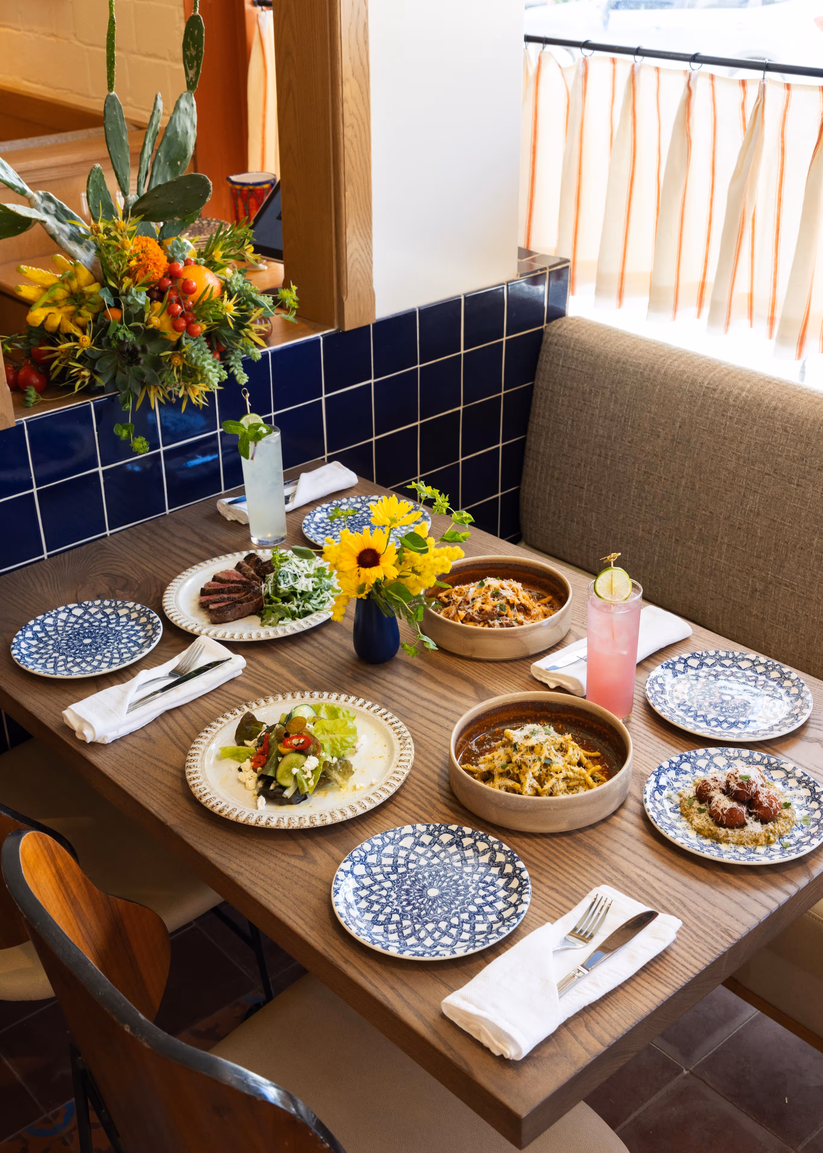 Wooden dining table set with four patterned plates, two bowls of pasta, a steak dish, a salad, two tall drinks, and a small vase with yellow flowers in a cozy restaurant corner.