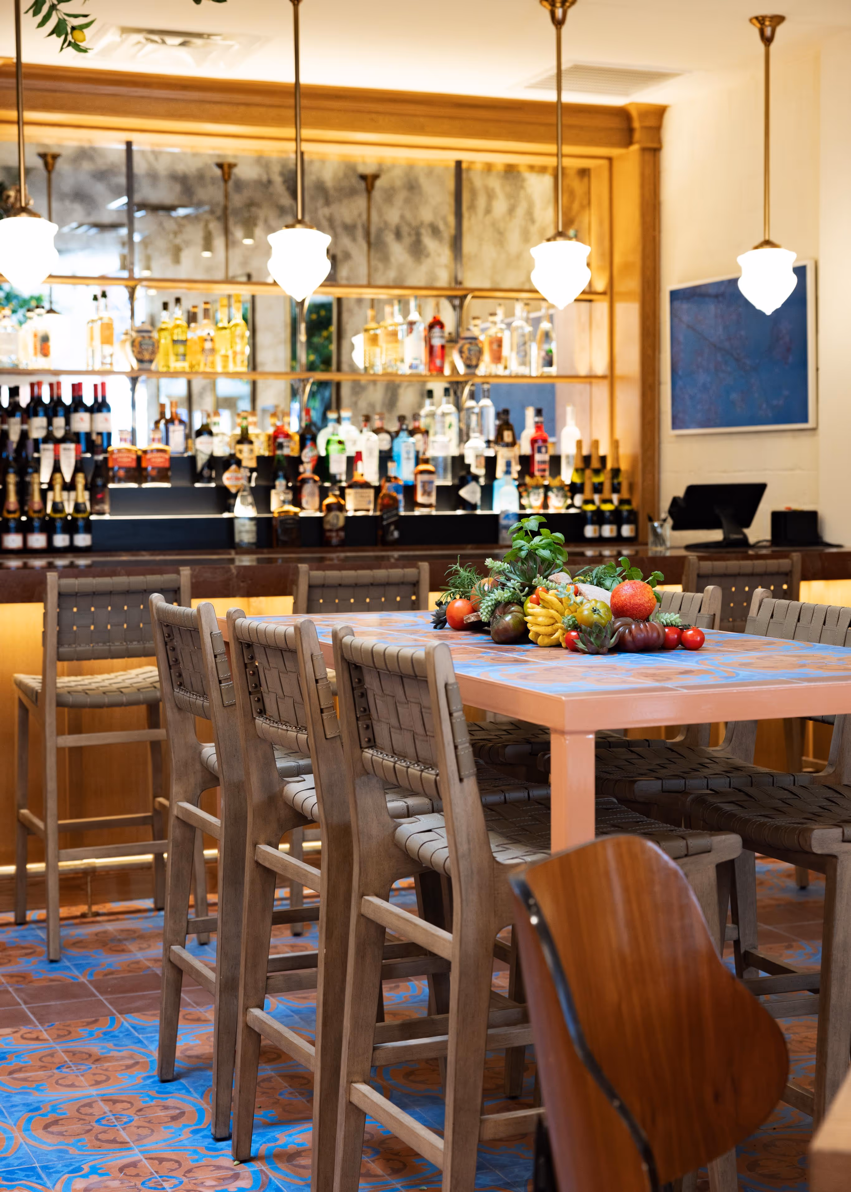Bar area with a tall table surrounded by woven seat chairs and a centerpiece of assorted fruits and herbs, illuminated by pendant lights.