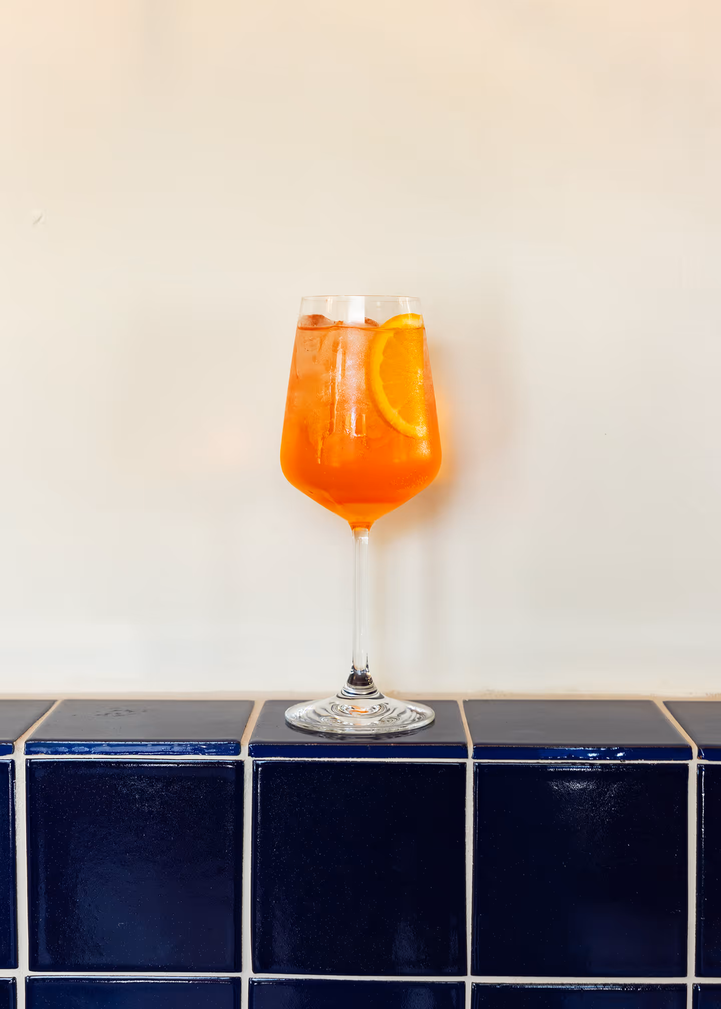 Glass of orange cocktail with ice and an orange slice on a blue tiled surface against a cream wall.