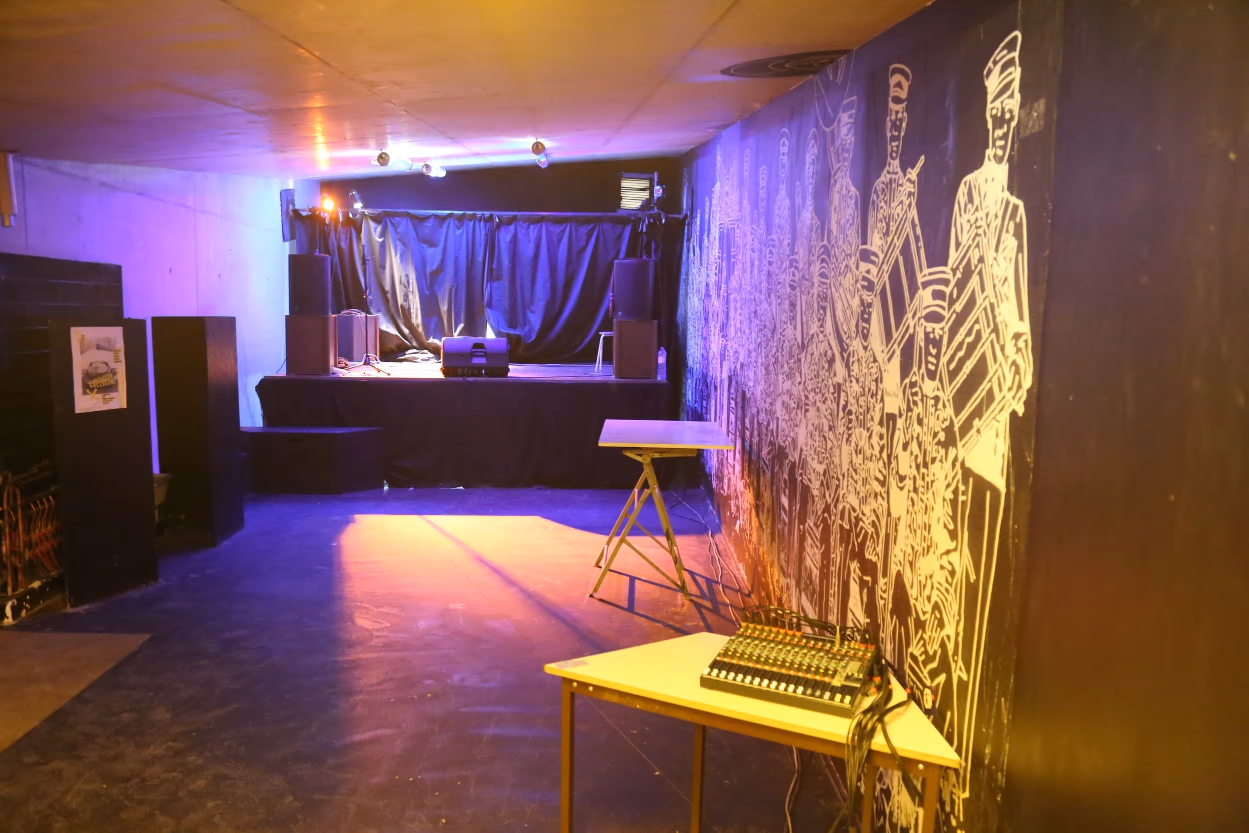 Salle de concert avec petite scène, éclairage coloré et mélangeur audio sur une table près d'un mur décoré de dessins en blanc.