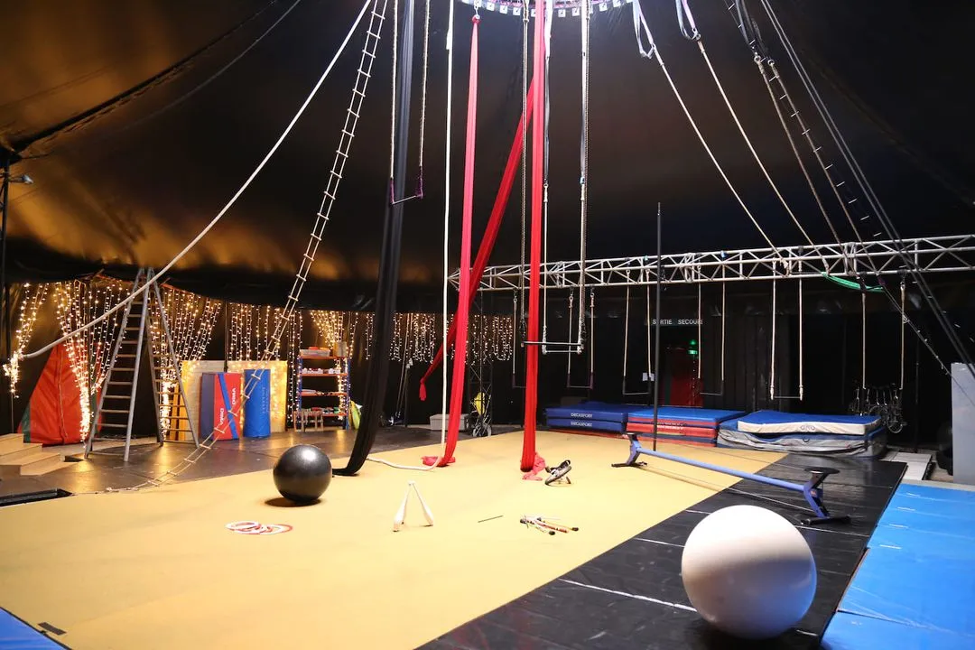 Intérieur d'un chapiteau de cirque équipé avec agrès suspendus, balles, massues et tapis de gymnastique.