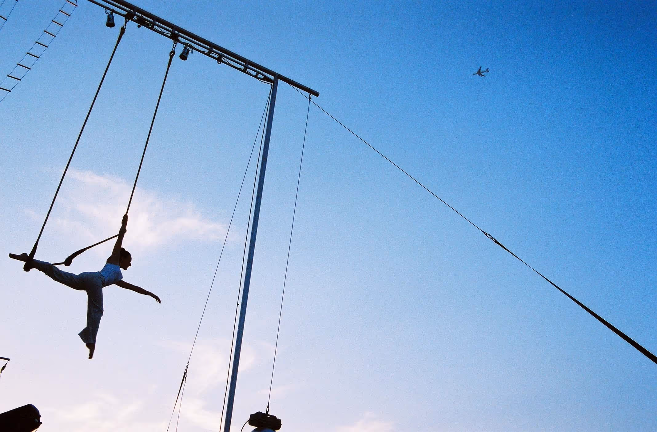Acrobate en plein air effectuant une pose élégante sur un trapèze contre un ciel bleu clair avec un avion en arrière-plan.