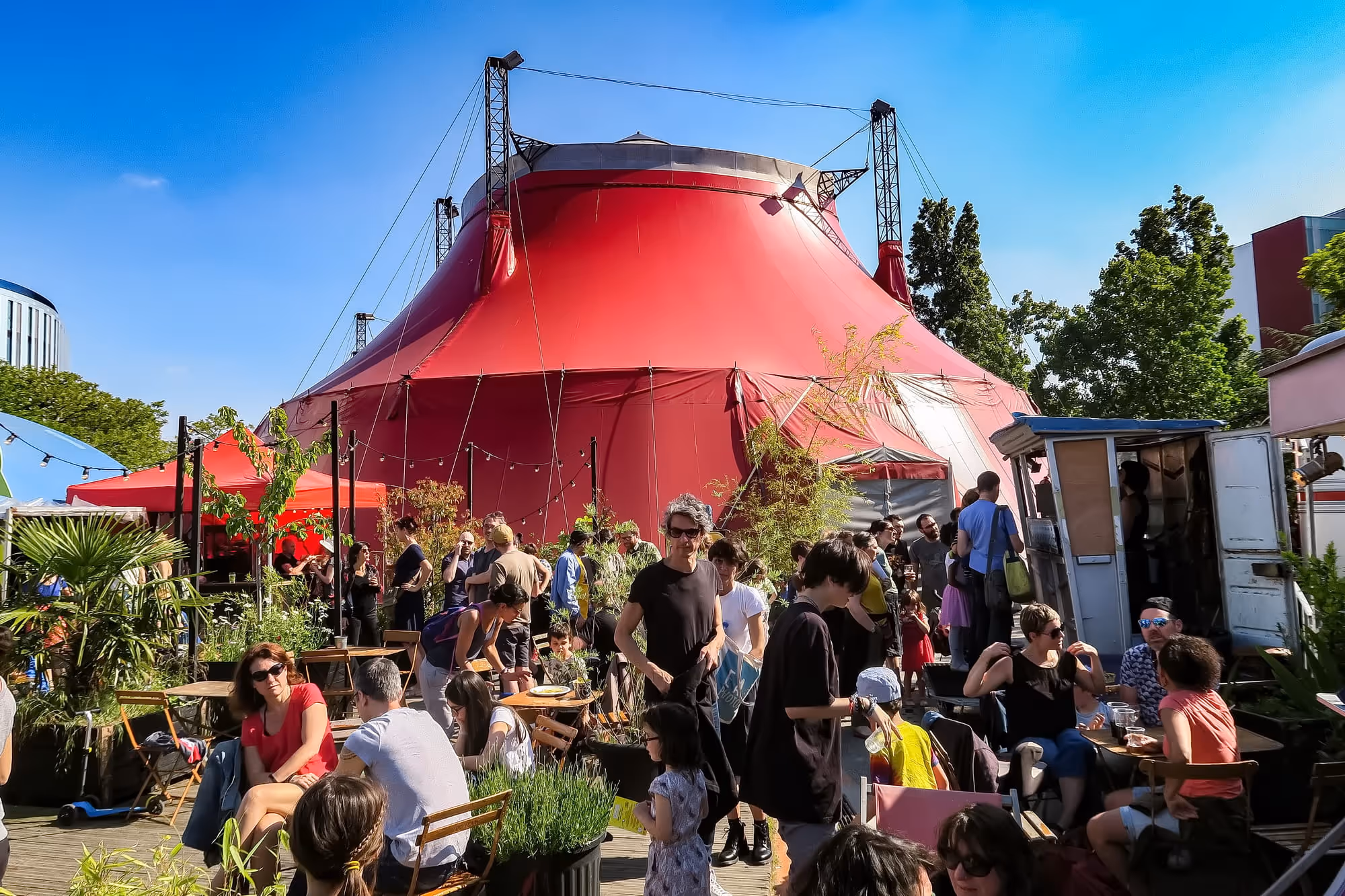 Chapiteau rouge du Cirque Électrique avec terrasse animée et architecture moderne en arrière-plan.